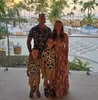 Family of four posing outdoors near water with palm trees and cityscape in the background.