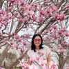 Woman with glasses and long dark hair standing amid pink cherry blossom trees in full bloom.